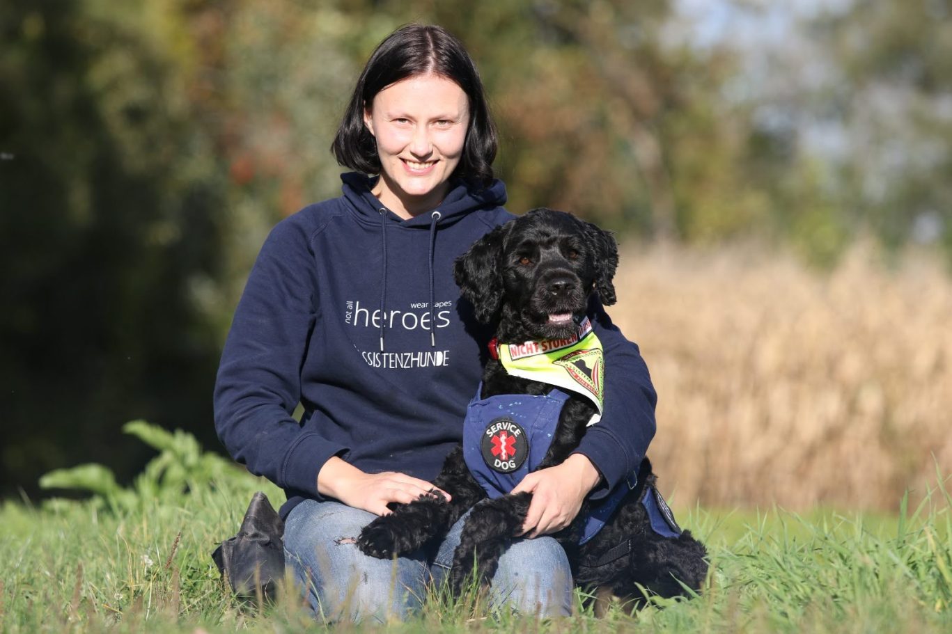 Frau mit mittellangen schwarzen Haaren kniet im Gras, auf ihrem Schoß liegt ihr gekennzeichneter Assistenzhund (portugiesischer Wasserhund)
